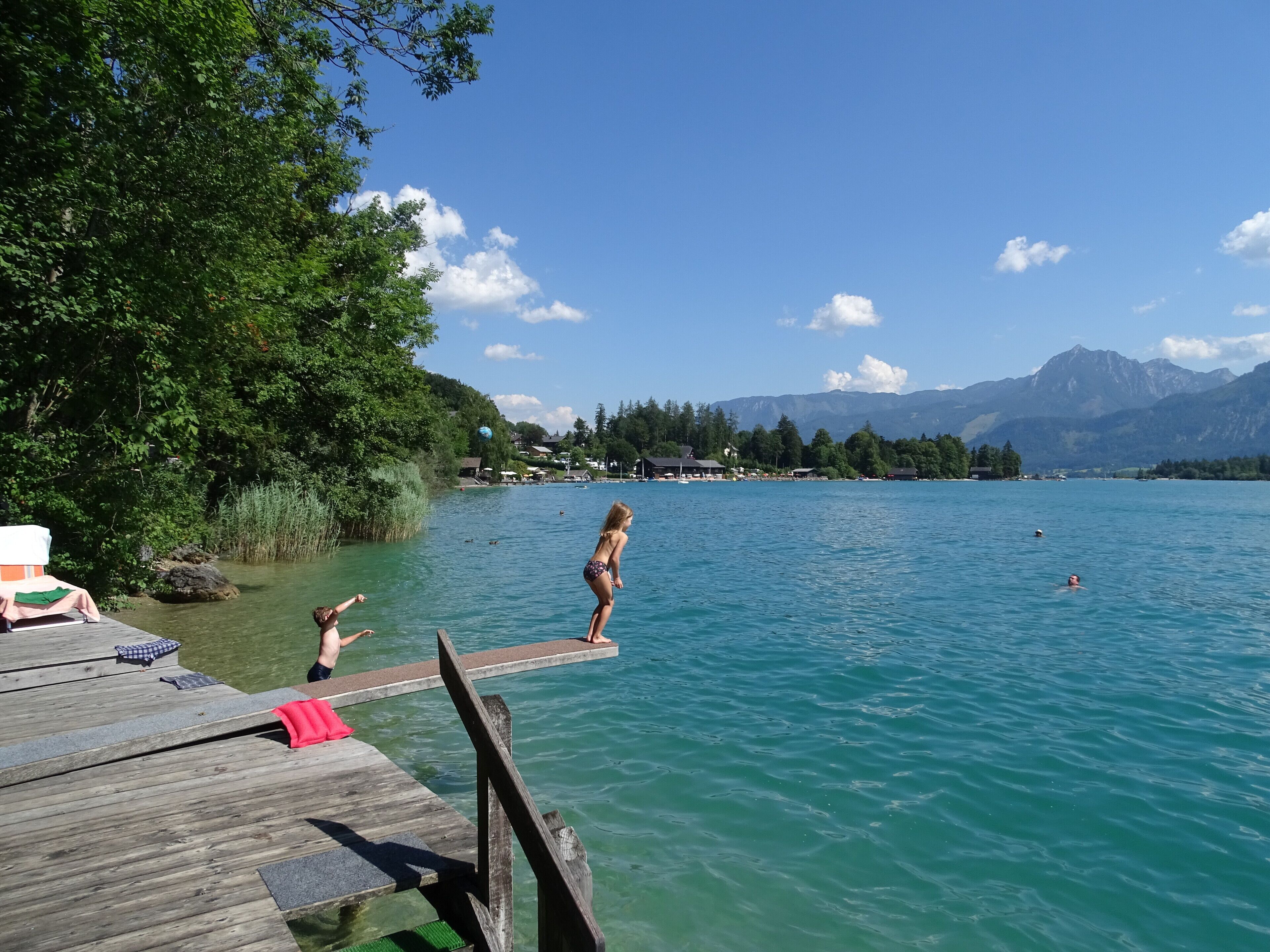 Eine Holzstege, der sich in einen See mit klarem Wasser erstreckt, umgeben von üppiger Vegetation und Bergen im Hintergrund.