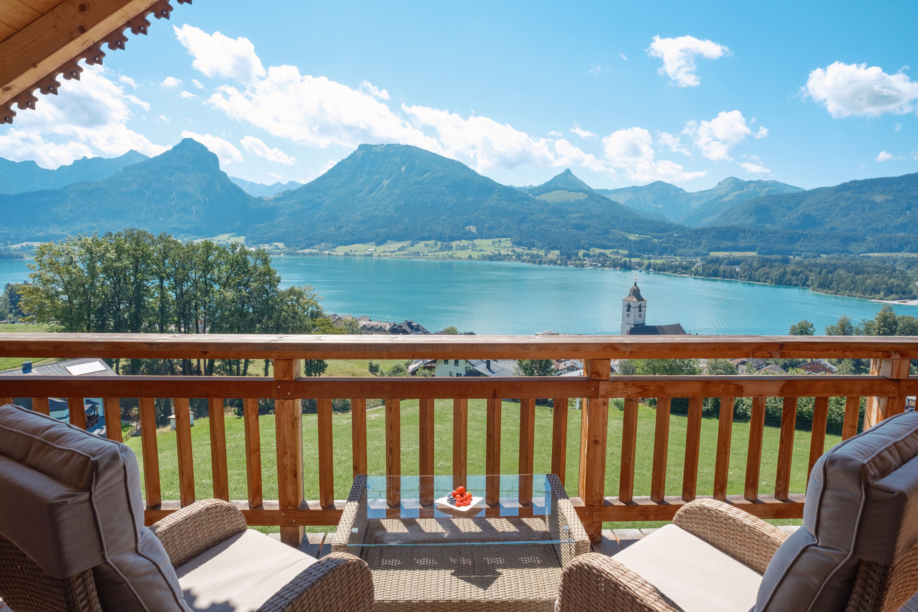 A balcony with wicker furniture overlooking a lake and mountains.