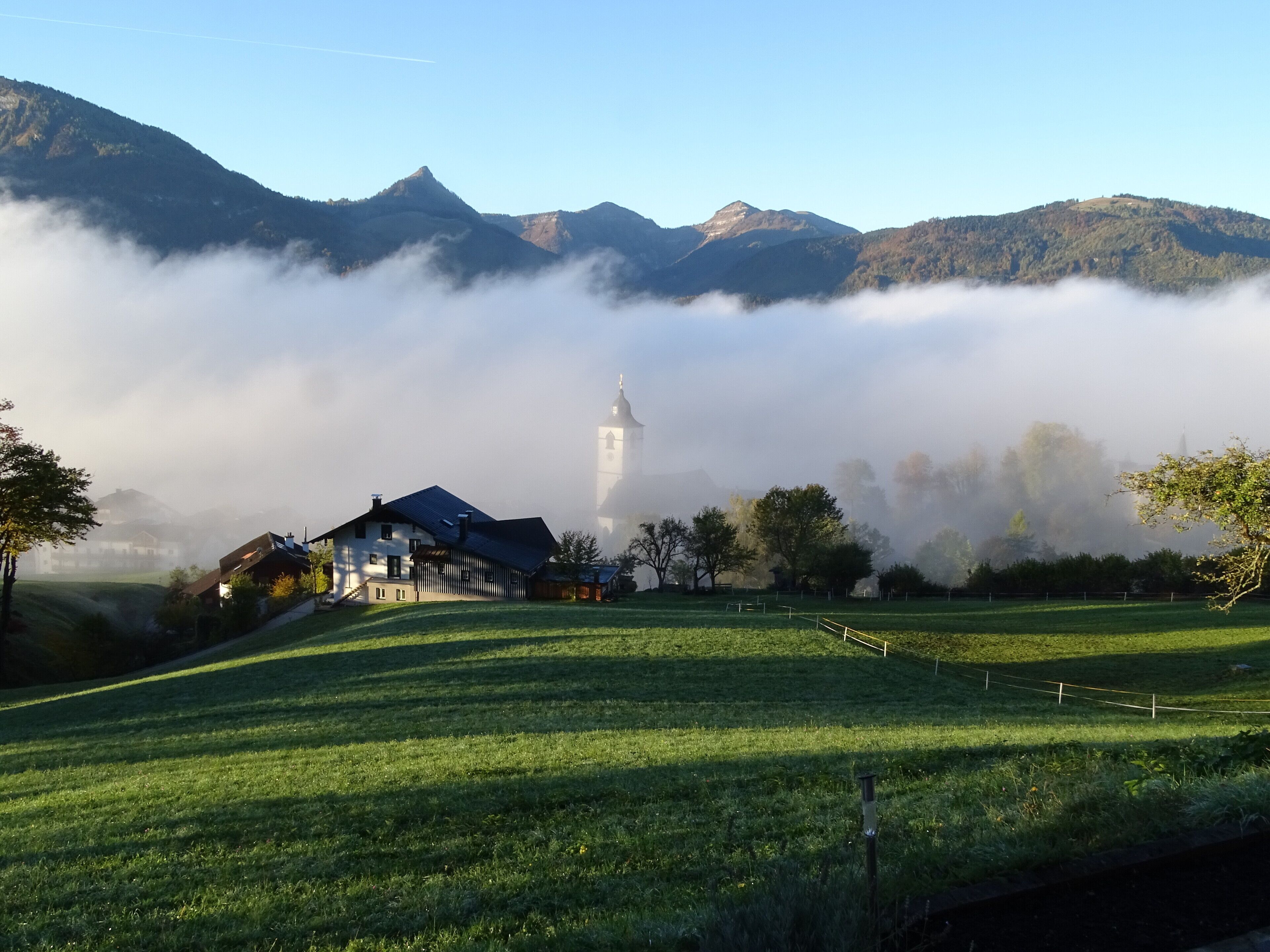 Paisaje rural con una torre de iglesia, una casa y montañas al fondo.