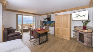 A room with a bed, a dining table with chairs, a kitchen area, a wooden wardrobe, a fireplace, and a balcony with a view of mountains. - Altroiterhof (Sankt Wolfgang im Salzkammergut)