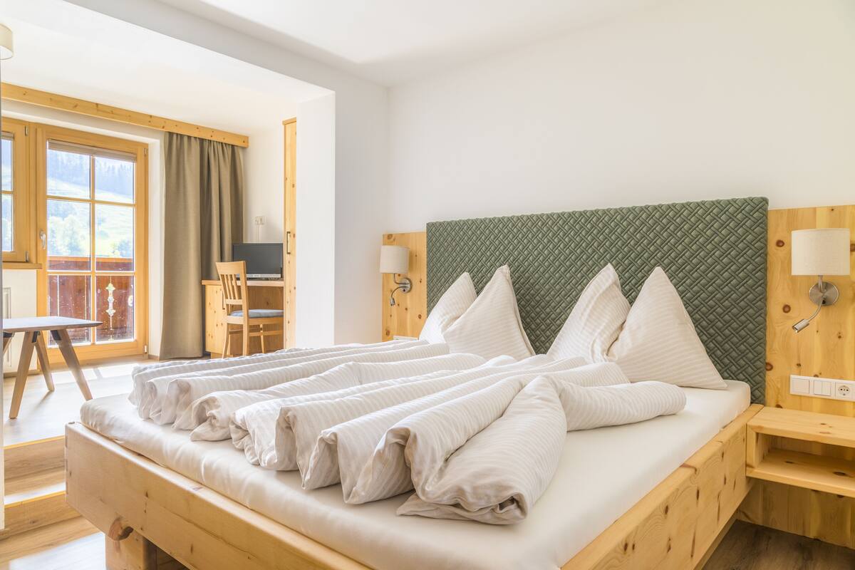 A neatly made bed with white linens, a wooden headboard, and a view of a balcony with a chair and table.