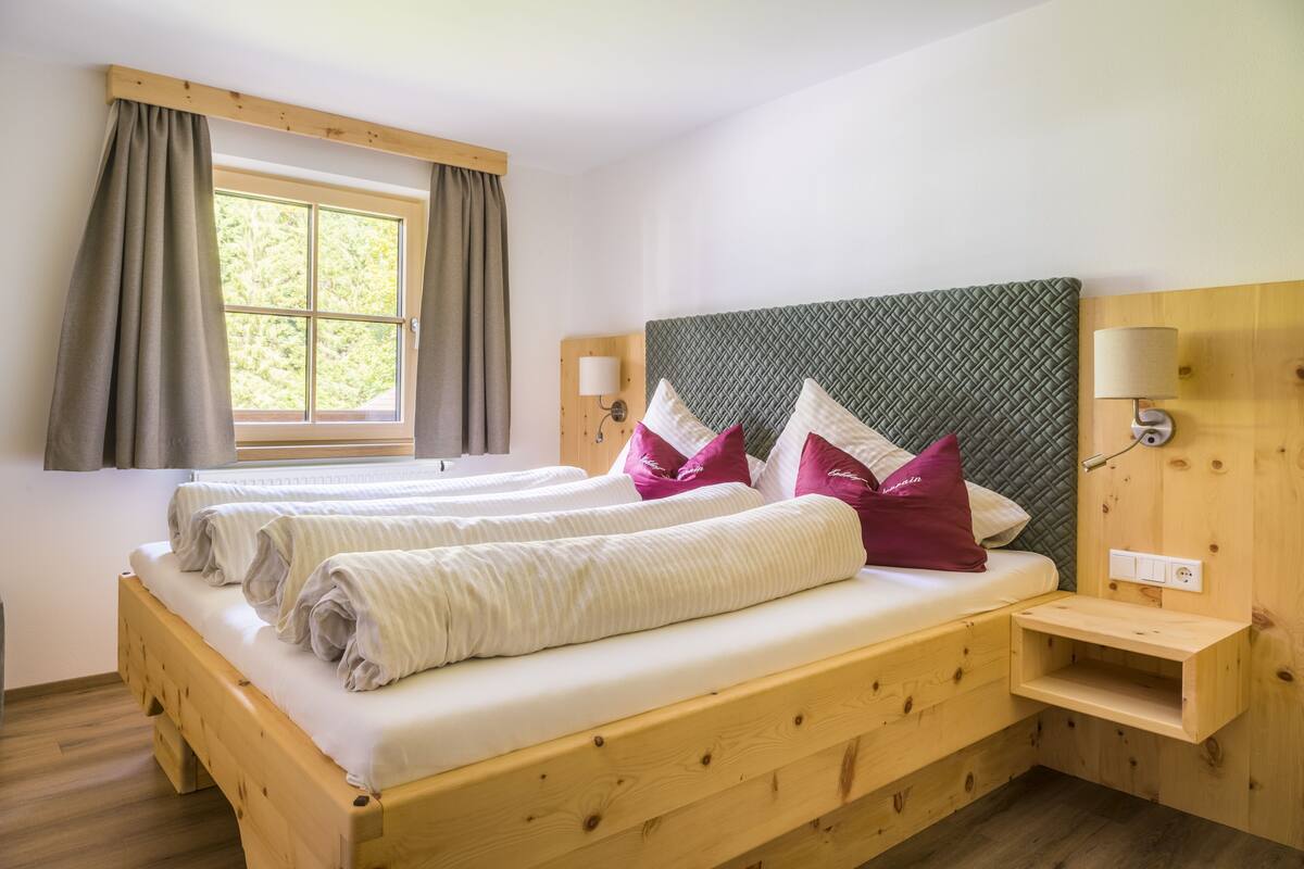 A double bed with two pillows, a wooden headboard, a bedside table, and a window with curtains.