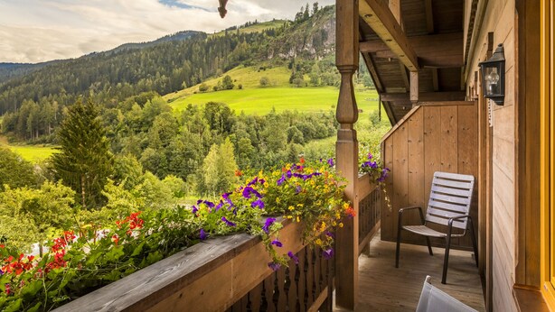 A wooden balcony with a chair and hanging flower baskets, overlooking a green valley and forested hills. - Erbhof Hinterrain (Leogang)