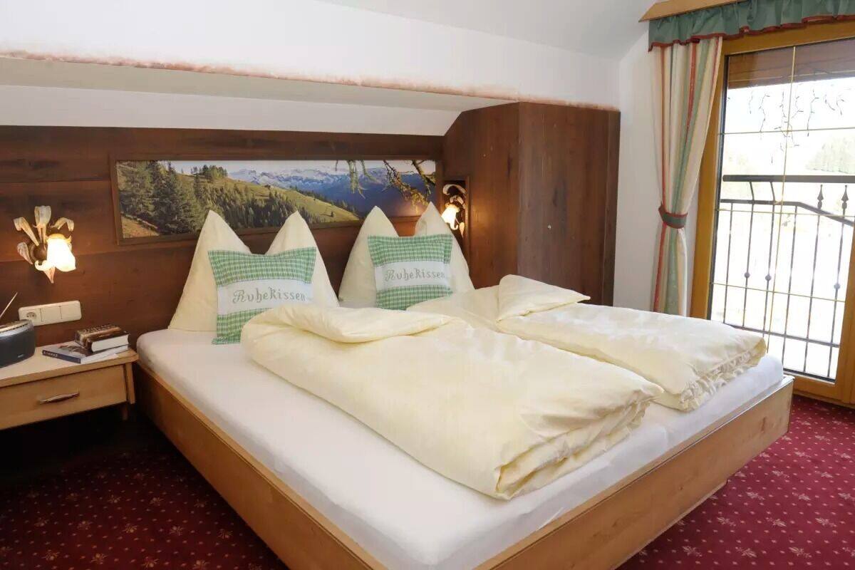 A neatly made bed with pillows labeled "Rubekissen" in a room with a wooden headboard, a nightstand with books, and a window with curtains.