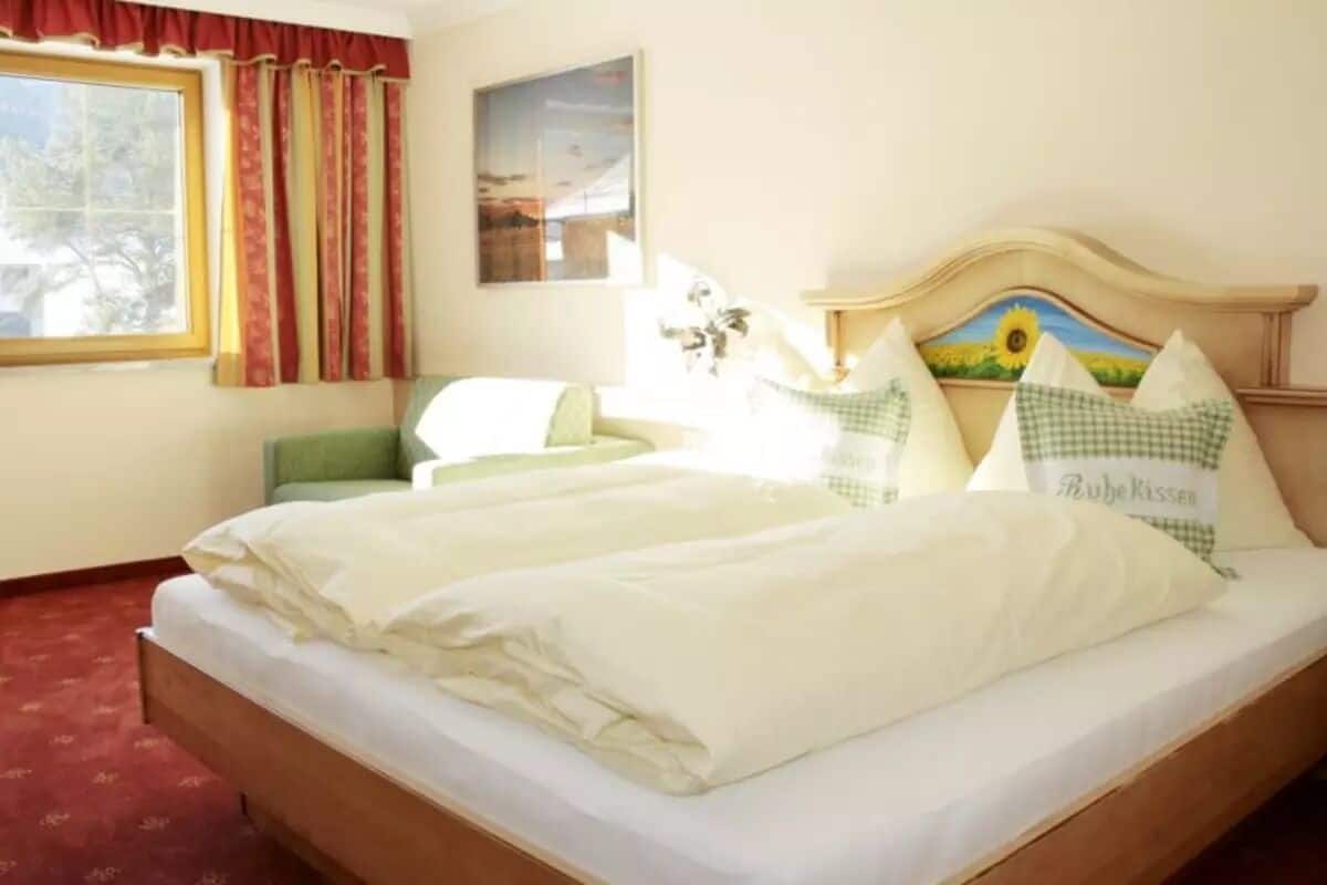 A double bed with white bedding, a green chair, a window with red curtains, and a painting on the wall.