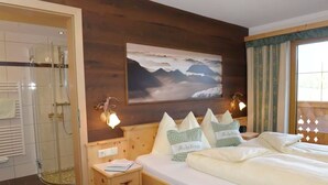 Habitació d'hotel amb un llit, una tauleta de nit, un quadre de muntanyes, una dutxa i una finestra amb cortines.