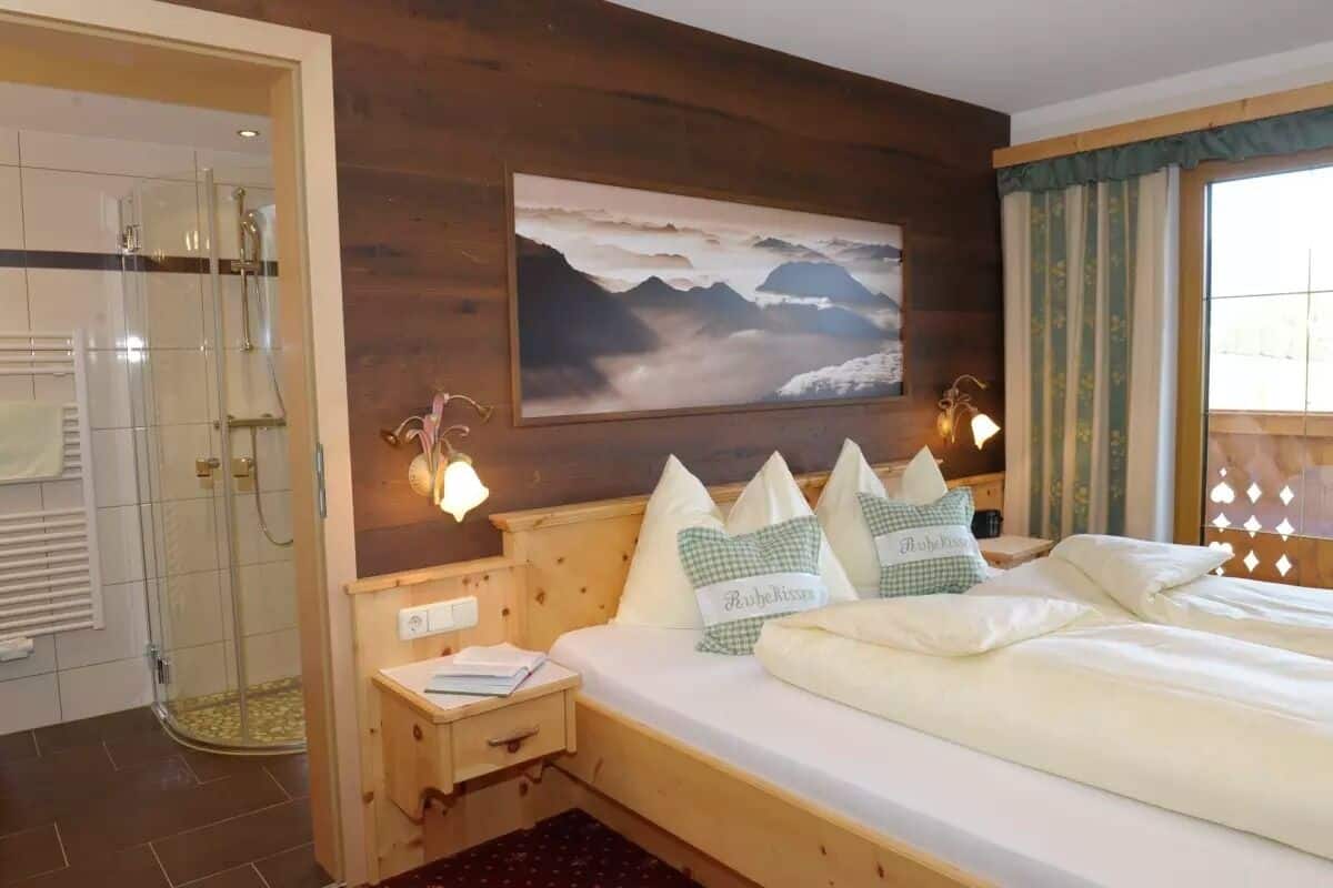 A hotel room with a bed, a nightstand, a painting of mountains, a shower, and a window with curtains.