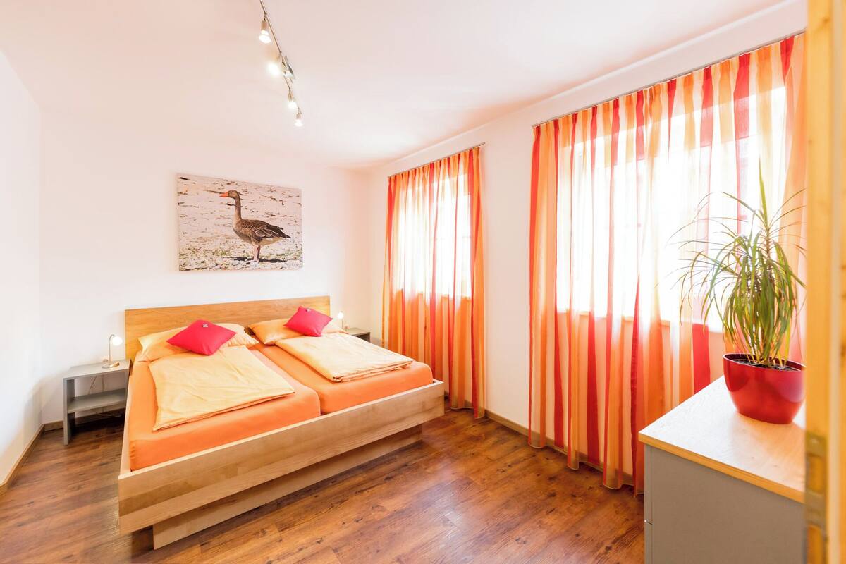 Una habitación con una cama de madera, ropa de cama naranja, una planta en maceta y un cuadro de un pato en la pared.