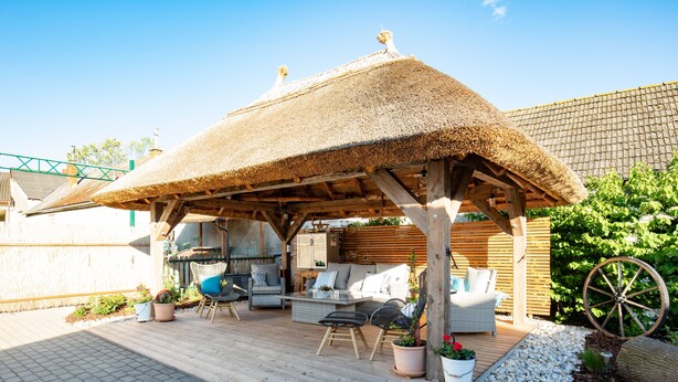 A covered outdoor seating area with wooden furniture, a thatched roof, and potted plants.