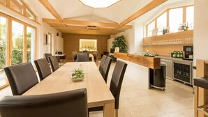 A spacious dining area with a long wooden table and black chairs, a kitchen with a wooden countertop and various appliances, and large windows allowing natural light. - Familie Bauer – Bioweingut Bauer (Großriedenthal)