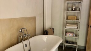 Een badkamer met een vrijstaand bad, een radiator, een plankje met toiletartikelen en een betegelde vloer.