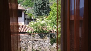 A view through a window with a curtain, featuring a stone wall, a tiled roof building, and a green hill in the background.