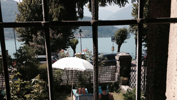 A view through a metal fence with a stone patio, a white umbrella, and a person sitting on a chair.