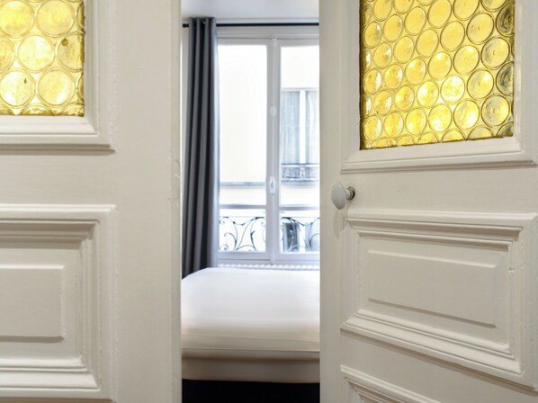 A hotel room with a bed, a window with a view of a balcony, and a door with a decorative glass panel.