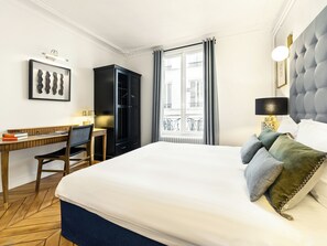 Een hotelkamer met een groot bed, een bureau, een stoel, een kledingkast, een raam met gordijnen en uitzicht op gebouwen.