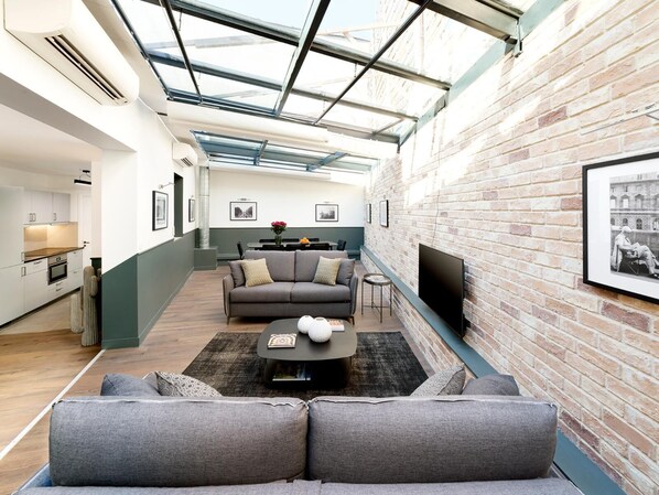 A modern living room with a glass ceiling, a sofa set, a coffee table, and a flat-screen TV mounted on the brick wall.