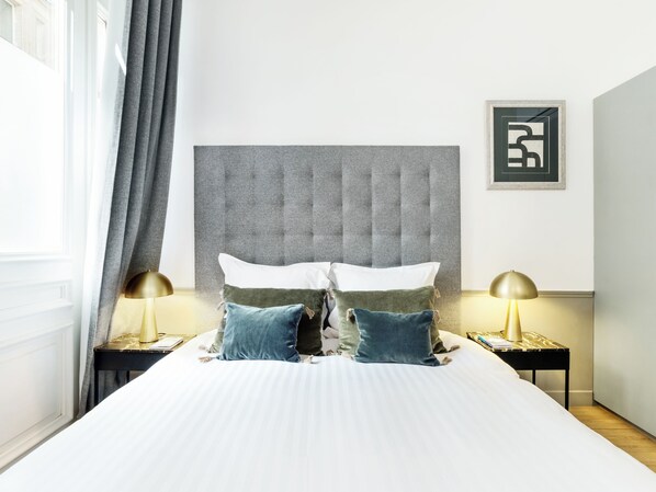 Une chambre d’hôtel avec un grand lit, deux tables de chevet, une tête de lit rembourrée de couleur grise et un tableau encadré au mur.