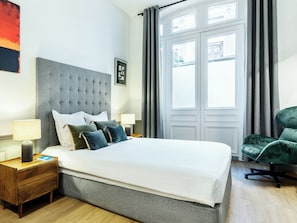 A hotel room with a large bed, two bedside tables, a green armchair, and a window with curtains.