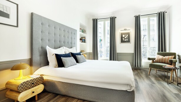 Quarto de hotel moderno com uma cama grande, uma mesinha de cabeceira, uma cadeira e vista para o exterior.