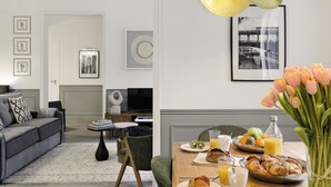A modern living room with a grey sofa, a coffee table, and a dining area with breakfast items. - LivinParis | Luxury AC 4 Bedrooms Opera I (Paris)