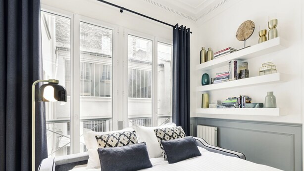 A modern bedroom with a large bed, a floor lamp, and a view of buildings outside the window. - LivinParis | Luxury AC 4 Bedrooms Opera I (Paris)