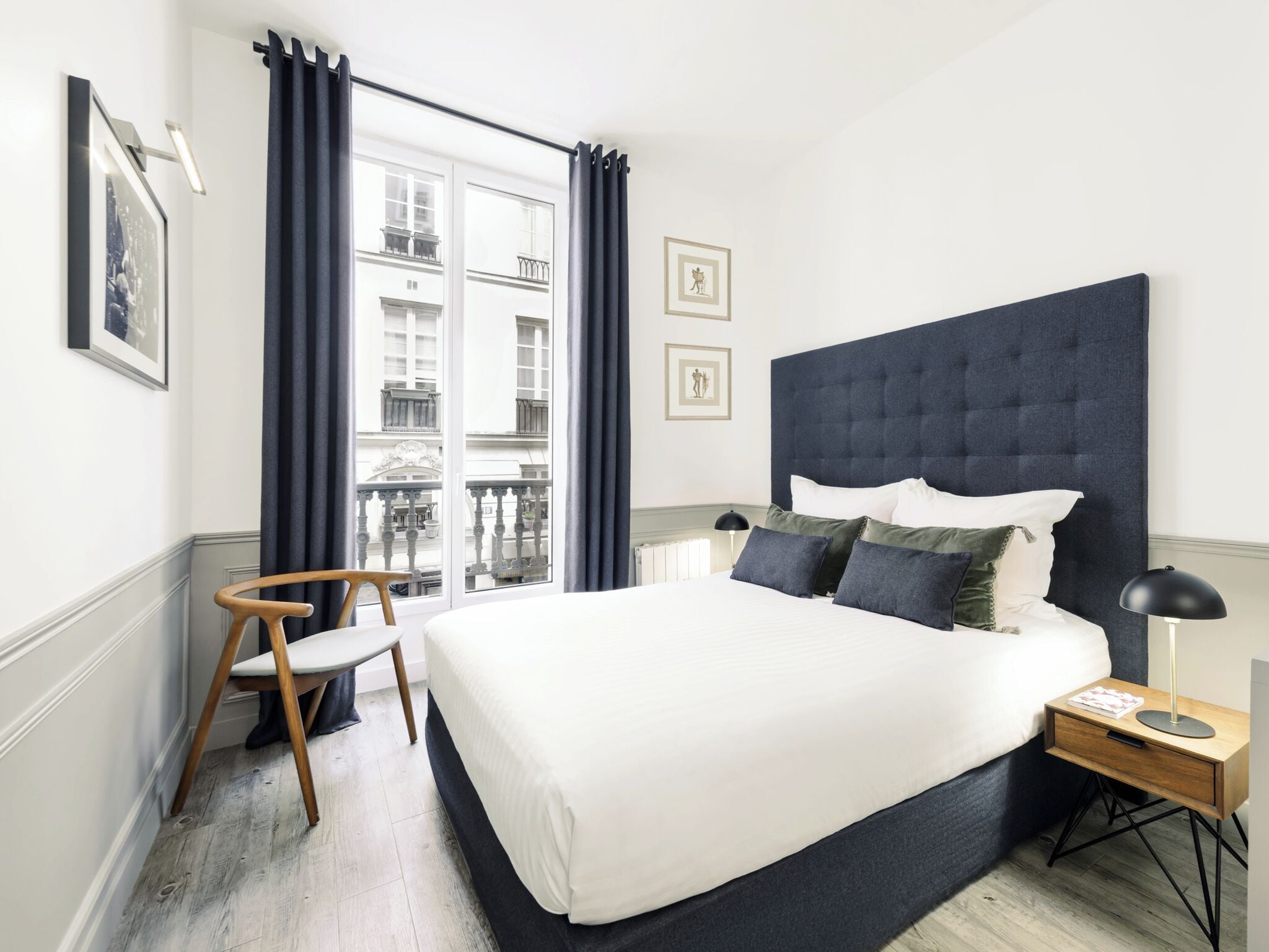 Una camera da letto con un letto grande, una sedia di legno, un comodino e vista sugli edifici attraverso la finestra.