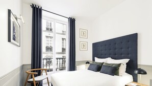 A bedroom with a large bed, a wooden chair, a nightstand, and a view of buildings through the window. - LivinParis | Luxury AC 4 Bedrooms Opera I (Paris)