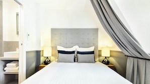 A hotel room with a bed, two bedside lamps, a headboard, a mirror, and a bathroom visible through an open door. - LivinParis | Luxury AC 3 Bedrooms Le Louvre - Eiffel View (Paris)