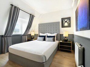 Habitación de hotel con una cama grande, dos mesitas de noche con lámparas, un radiador y una ventana con cortinas.