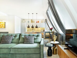 A modern living room with a green sofa, a coffee table, and a TV. There is a kitchen in the background with a dining area.