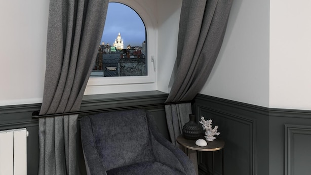 A corner room with a view of a cityscape through a window, a grey armchair, and a small round table with decorative items. - LivinParis | Luxury AC 2 Bedrooms Le Louvre - Sacré Cœur View (Paris)
