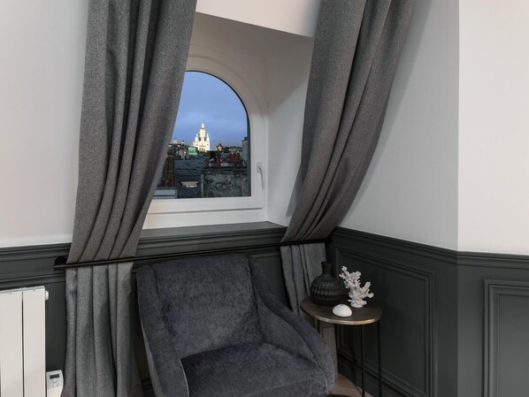 Habitación de esquina con vista a un paisaje urbano a través de una ventana, un sillón gris y una mesita redonda con objetos decorativos.