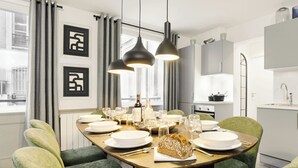 A modern dining room with a wooden table set for six, green upholstered chairs, pendant lights, and a view of the city through large windows.