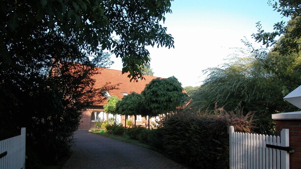 A paved pathway leading to a building with a red roof, surrounded by greenery and a white picket fence. - Small vacation apartment at Wulfkamp (Velen)