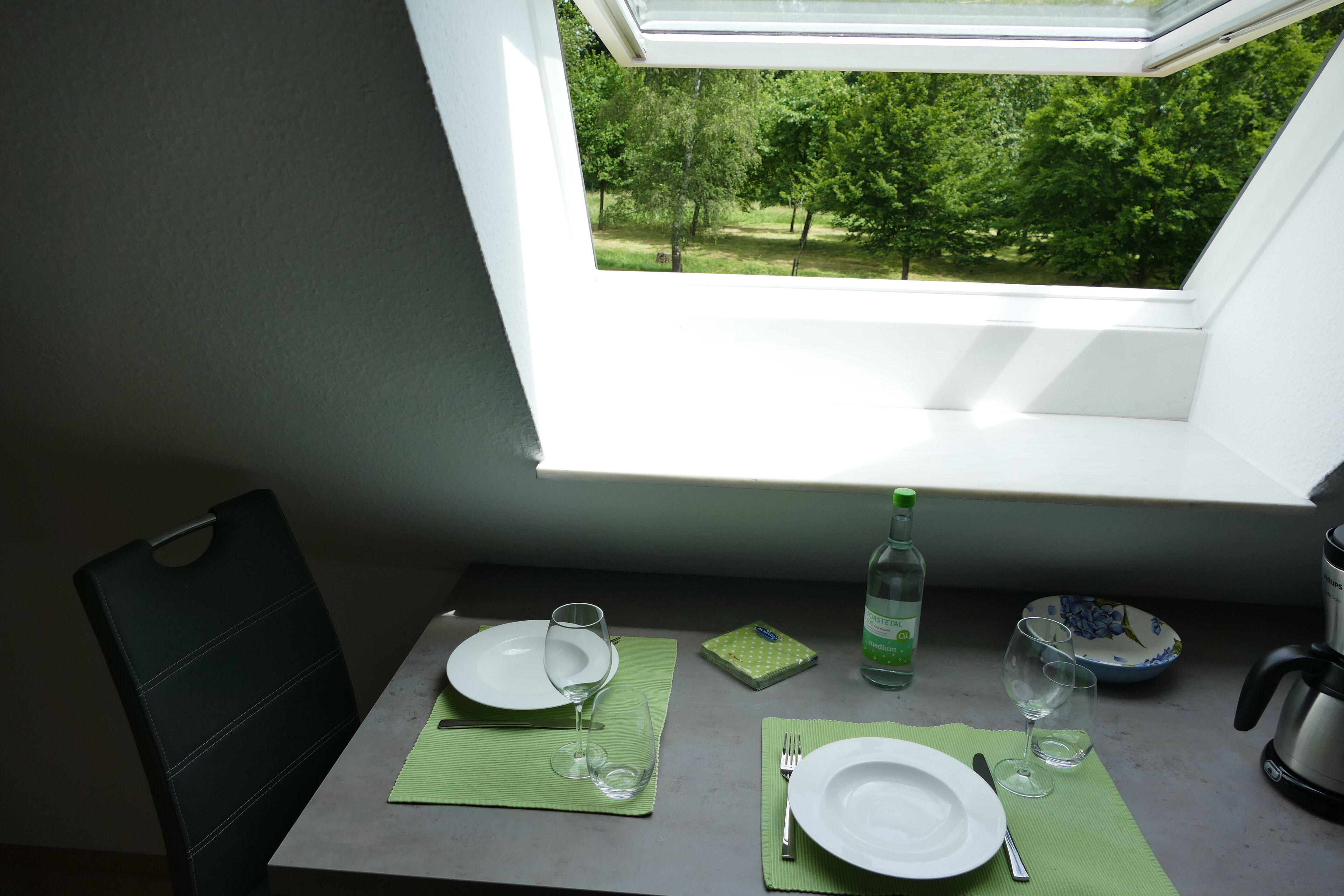 Une table à manger donnant sur des arbres, vue à travers une fenêtre, une bouteille d’eau et une bouilloire.