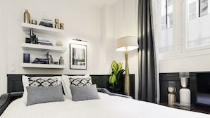 A modern hotel room with a large bed, a bedside table with a lamp, a bookshelf, and a framed picture on the wall. - LivinParis | Luxury AC 3 Bedrooms République I (Paris)