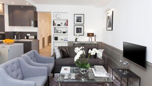 Een moderne woonkamer met een grijze bank, een glazen salontafel en een televisie. Achtergrond is een keuken met donkere kastjes en een fruitschaal.