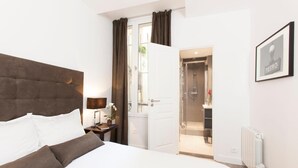 Een moderne slaapkamer met een groot bed, een nachtkastje met een lamp, een spiegel en een deur naar een badkamer met een douche.