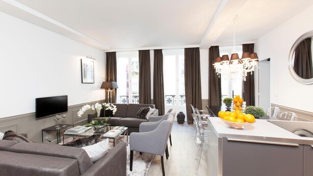 Een moderne woonkamer met een bank, salontafel en televisie. Er is een eethoek met een tafel en stoelen, en een keuken met een fruitschaal.