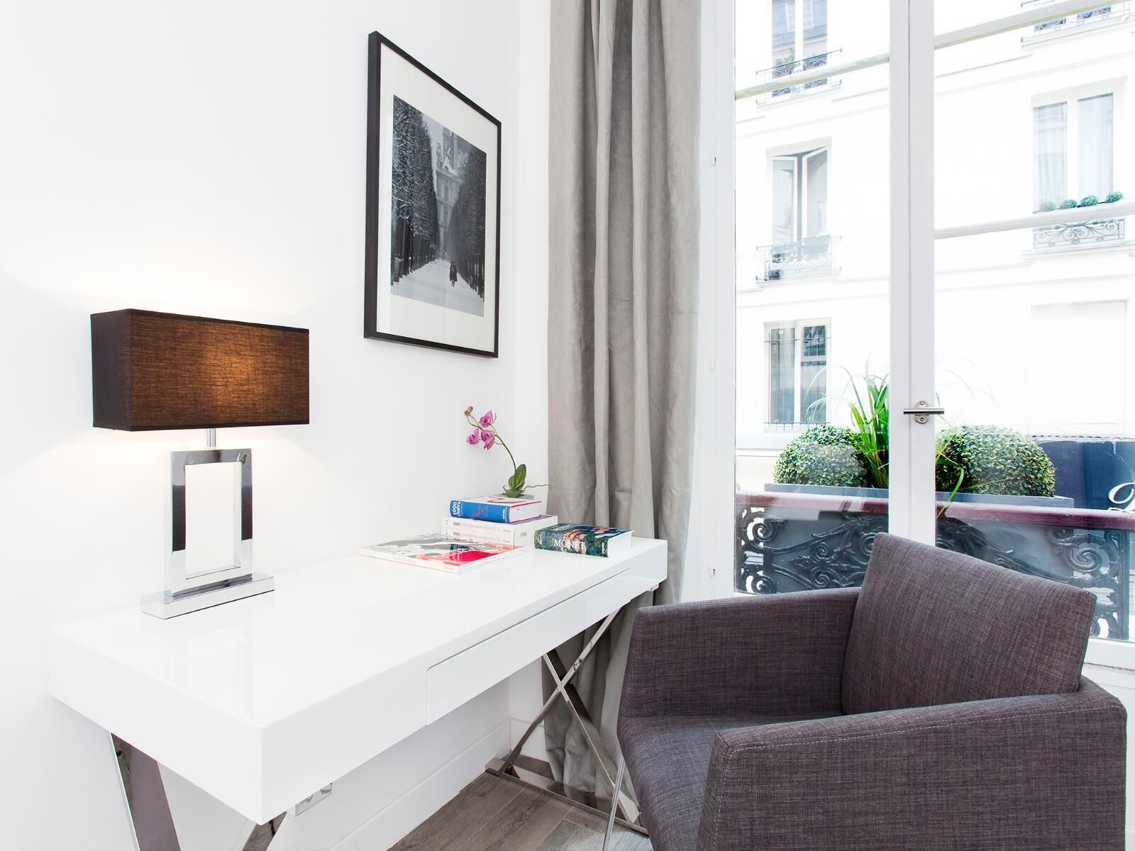 Ein modernes Zimmer mit einem weißen Schreibtisch, einem Schwarz-Weiß-Foto in einem Rahmen, einer Lampe und Blick auf einen Balkon mit Pflanzen.