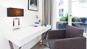 Ein modernes Zimmer mit einem weißen Schreibtisch, einem Schwarz-Weiß-Foto in einem Rahmen, einer Lampe und Blick auf einen Balkon mit Pflanzen.