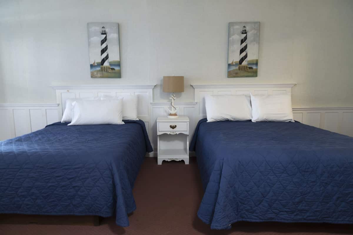 Ein Zimmer mit zwei Betten, jedes mit einer blauen gesteppten Bettdecke und weißen Kissen. Dazwischen steht ein weißer Nachttisch mit einer Lampe. An der Wand über den Betten hängen zwei Bilder von Leuchttürmen.