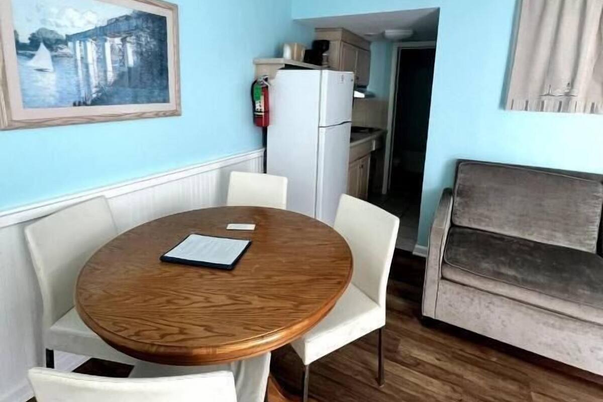 Ein gemütlicher Raum mit einem runden Holztisch, weißen Stühlen, einem grauen Sofa, einer kleinen Küchenzeile und einem gerahmten Bild an der Wand.