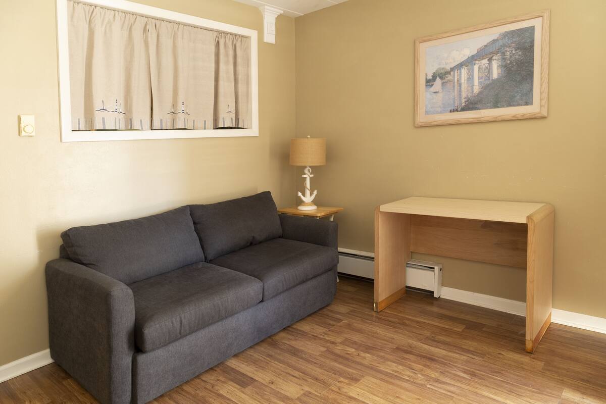 Ein Zimmer mit grauem Sofa, einem Holztisch mit Lampe, einem Gemälde an der Wand und einem Fenster mit Vorhängen.
