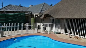 Una piscina con cerca blanca, una estructura de techo de paja y un patio de ladrillo.
