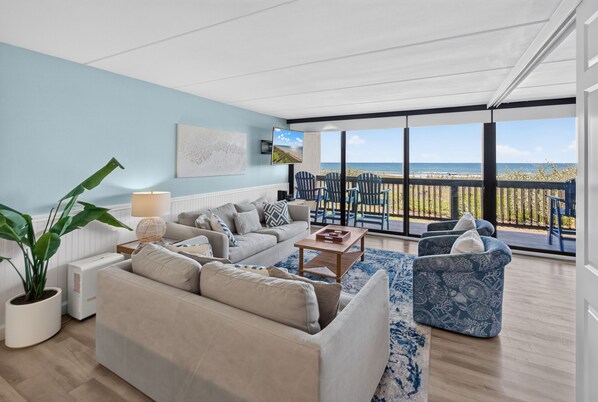 A living room with a sofa, coffee table, and chairs, a large potted plant, and a view of the ocean through a balcony.