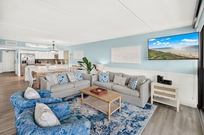 Un salon moderne avec un canapé gris, des coussins à motifs bleus, une table basse en bois et un grand écran de télévision diffusant une scène de plage.