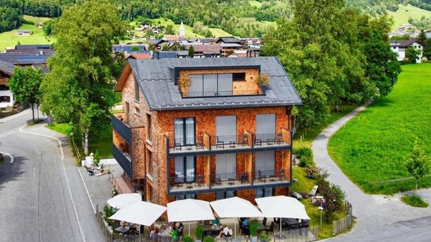 A two-story brick building with a balcony, surrounded by lush greenery and a mountain backdrop.