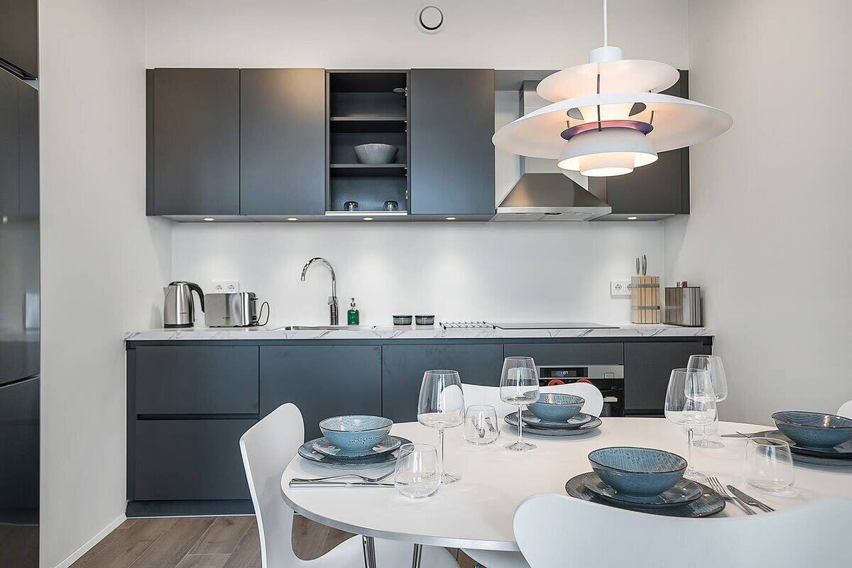 Una cucina moderna con mobili scuri, un tavolo da pranzo bianco apparecchiato con ciotole blu e bicchieri da vino, e una lampada a sospensione sopra.