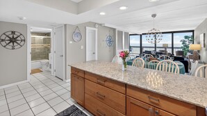 A modern kitchen with a large island, white tiled flooring, and a dining area with a view. - Gulf Shores 506 Fifth Floor Beachfront View 2 Bedroom 2 Bathroom Condo (Port Aransas)